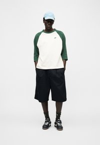 Νέος άνδρας που φοράει λευκό και πράσινο μπλουζάκι baseball της Nike, μαύρα σορτς, μαύρα αθλητικά παπούτσια Nike, σκουροπράσινες κάλτσες και ανοιχτό γαλάζιο καπέλο Nike, όρθιος σε χαλαρή στάση.