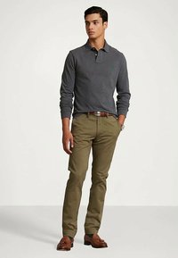 Mężczyzna w ciemnoszarej koszulowej bluzie z długim rękawem, w oliwkowych spodniach, z brązowym skórzanym paskiem i brązowymi mokasynami z frędzlami, stojący z jedną ręką w kieszeni.