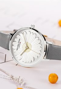 Rund klocka med en texturerad vit blommig urtavla, silverdetaljer och gyllene timmarkörer. Grå läderrem. Har en enkel krona.