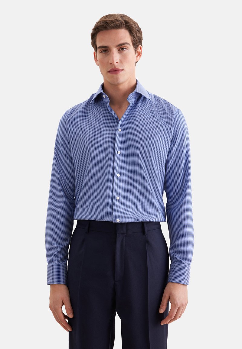 Jeune homme portant une chemise à motifs bleue à manches longues et un pantalon bleu marine foncé, debout devant un fond clair uni.