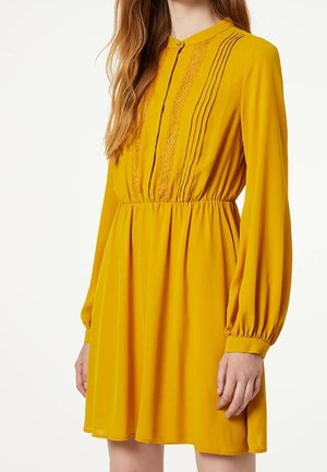 Robe longue à manches longues jaune avec taille froncée, détails en dentelle et plis nervurés sur la poitrine, col boutonné et manches amples avec poignets.