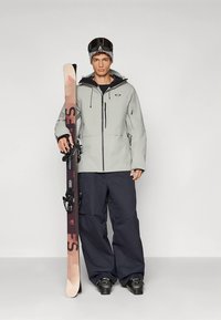 Ski-Outfit mit einer hellgrauen wasserdichten Jacke mit hohem Kragen, kombiniert mit dunkelblauen Cargohosen und schwarzen Schneestiefeln. Ski in einer Hand gehalten.