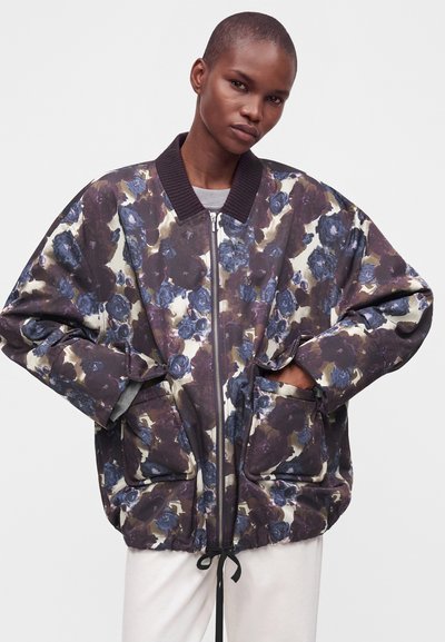 Personne aux cheveux courts portant une veste bomber ample à motif floral avec des tons bleus et marron et un pantalon blanc, les mains dans les poches de la veste.