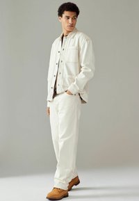 Chaqueta blanca de algodón con frente de botones, bolsillos laterales y costuras tonal, combinada con pantalones blancos a juego y zapatos beige.