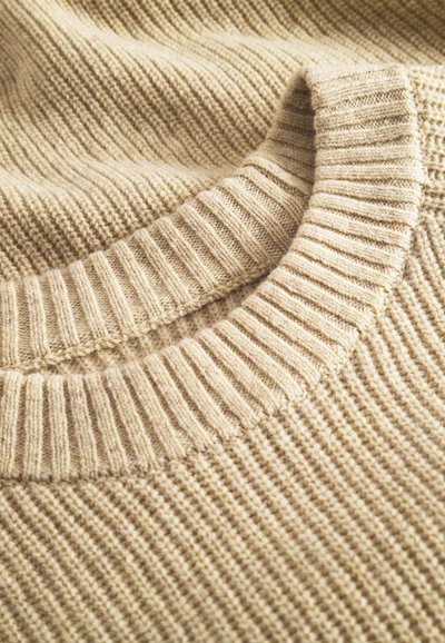 Bež pleteni pulover s rebrastim ovratnikom in teksturo, izdelan iz mehkega materiala z vidnimi šivi.