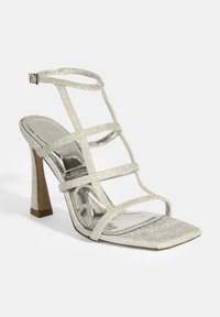 Glitzernde, silberne Sandalette mit hohen Absätzen und metallischen Akzenten. Verfügt über eine quadratische Zehenpartie und einen schlanken Stiletto-Absatz. Verstellbarer Knöchelriemen.