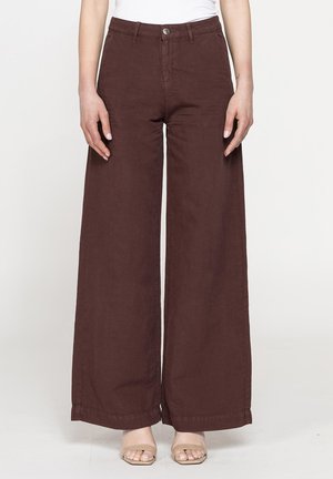 Donna che indossa pantaloni marroni a vita alta e gamba larga abbinati a sandali aperti color nudo, in piedi su uno sfondo chiaro uniforme.
