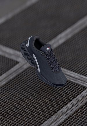 Baskets de sport noires avec amorti d'air visible sur une surface en grille métallique, montrant des textures et des lacets détaillés.