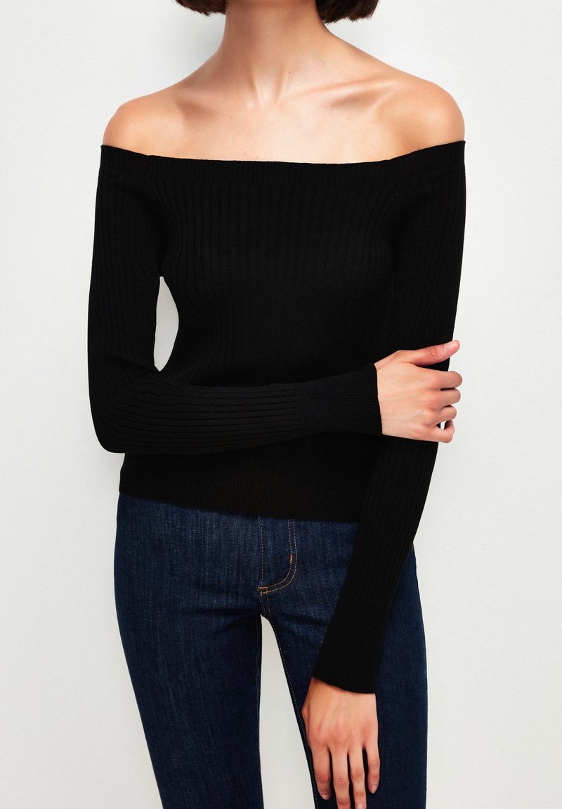 Femme portant un pull noir côtelé à épaules dénudées et un jean bleu foncé, debout avec un bras croisé et la main reposant sur l'autre bras.