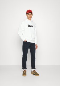 Biely mikina s čiernym logom "levi's", námornícke džínsy a olivovo zelené sneakers. Doplnky zahŕňajú červen�ú čiapku. Neutrálny pozadie.