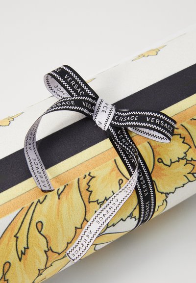 Dárkově zabalený předmět se stuhami značky Versace v černé a bílé barvě, zdobený dekorativním vzorem zlatých listů na bílém papíře.