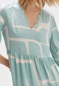 Vaaleansininen mekko, jossa on V-aukko, valkoisia geometrisia kuvioita, kerätty vyötärö ja pitkät hihat teksturoidusta kankaasta.