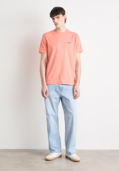 T-shirt corail à manches courtes avec un petit logo, associé à un jean droit bleu clair et des baskets blanches avec une semelle beige.