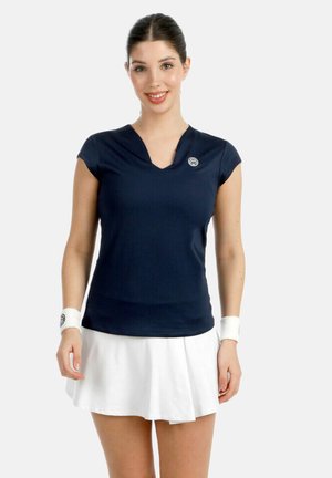 Frau trägt marineblaues Sportshirt mit V-Ausschnitt, weißen Faltenrock und passende Handgelenkbandagen, lächelt mit zurückgebundenem Haar vor weißem Hintergrund.