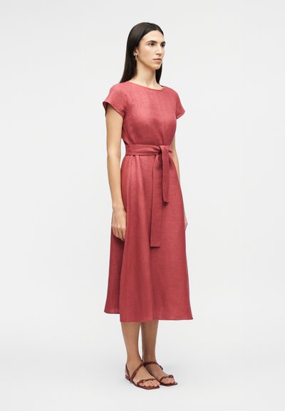 Γυναίκα με μακριά σκούρα μαλλιά που φοράει φόρεμα σε μήκος μέχρι το γόνατο σε σκούρο ροζ χρώμα, με κοντά μανίκια, ζώνη στη μέση και καφέ επίπεδα σανδάλια, στραμμένη στο πλάι.