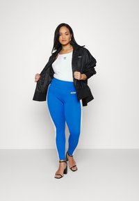 Črn bombnik, bela majica brez rokavov in modre legice z belimi stranskimi črtami. Črne sandale z visokimi petami dopolnjujejo športni videz.