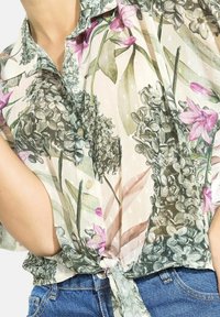 Floral button-up shirt featuring light pink base with green and purple flower patterns, short sleeves, and a tied hem detail.