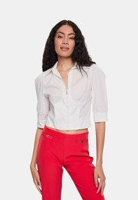 Blusa bianca corta con colletto, maniche a sbuffo corte e bottoni frontali, abbinata a pantaloni rossi a vita alta con dettagli dorati.