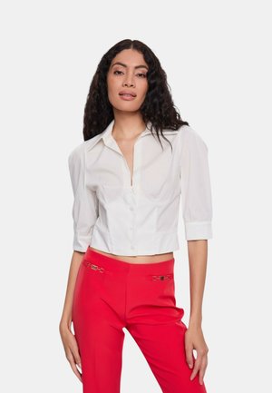 Blusa bianca corta con colletto, maniche a sbuffo corte e bottoni frontali, abbinata a pantaloni rossi a vita alta con dettagli dorati.
