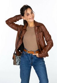 Veste en cuir synthétique marron avec un col de chemise, portée sur un haut rayé. Associée à un jean en denim taille haute et une ceinture marron.