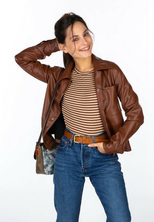 Veste en cuir synthétique marron avec un col de chemise, portée sur un haut rayé. Associée à un jean en denim taille haute et une ceinture marron.