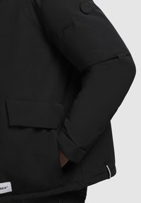 Chaqueta acolchada negra con diseño con cremallera, bolsillos laterales y una tela texturizada. Incluye un logo circular en la parte superior del brazo.