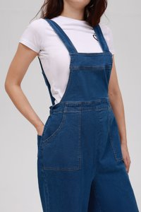 Μπλε τζιν φόρμα με ρυθμιζόμενους ιμάντες, μεγάλες μπροστινές τσέπες και άνετη εφαρμογή. Συνδυασμένη με ένα εφαρμοστό λευκό μπλουζάκι από κάτω.