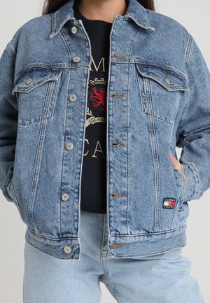 Persona que lleva una chaqueta vaquera azul claro sobre una camiseta negra con gráfico y vaqueros azul claro, con una mano en el bolsillo de la chaqueta.