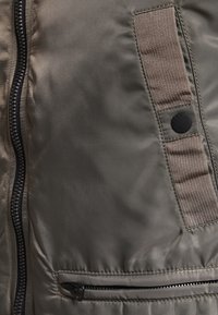 Polstrovaná vesta v tlumené zelené barvě, s černým zipem, texturovanou tkaninou a akcentem z plátna se zakulacenou nášivkou.