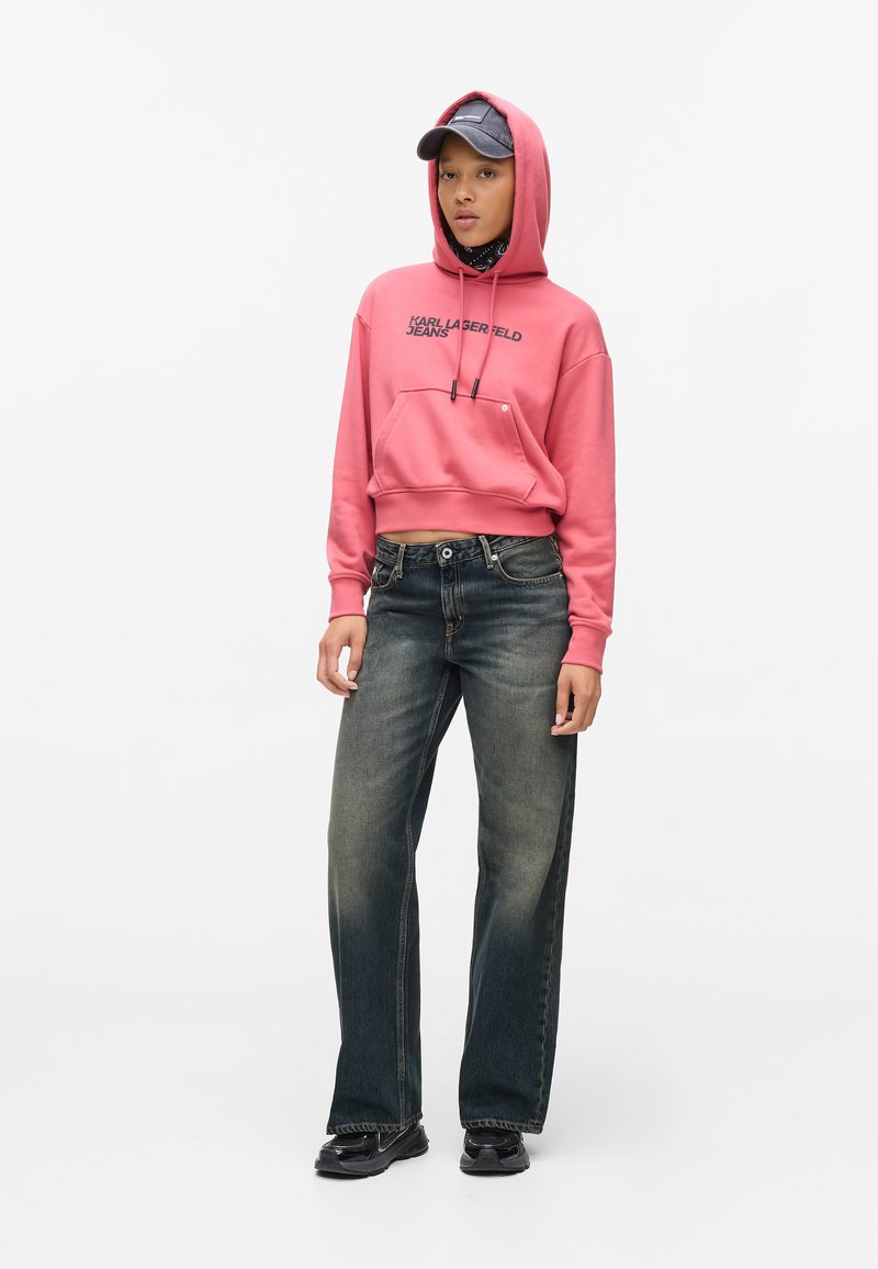 Rosa verkürzter Hoodie mit schwarzem Text, Fronttasche und Kordelzug; ausgebleichte blaue weitgeschnittene Jeans; schwarze Schuhe; dunkle Kappe mit Logo.