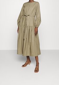 Donna che indossa un abito lungo, verde oliva, con bottoni e maniche lunghe, abbinato a sandali con tacco color beige.