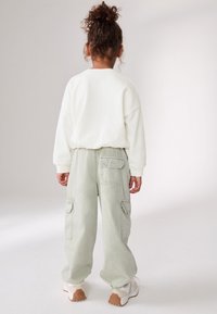Sweat-shirt blanc à coupe ample, associé à un pantalon cargo vert clair avec poches latérales, complété par des baskets blanches et beiges.