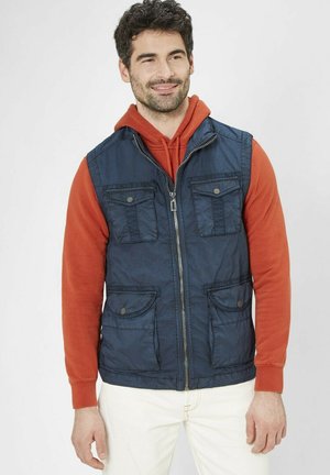 Mann mit kurzen lockigen Haaren, der eine marineblaue Zip-up-Weste über einem orangen Kapuzenpullover und weißen Hosen trägt, leicht lächelnd, vor weißem Hintergrund.