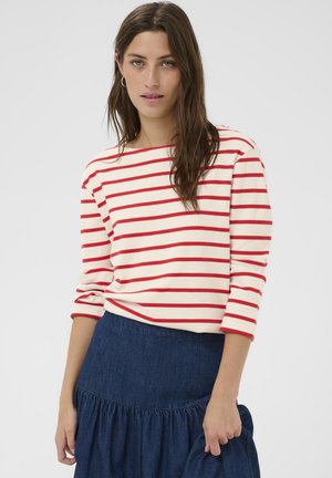 Vrouw met lang bruin haar, een rood-wit gestreept shirt met lange mouwen en een blauwe spijkerrok, staand tegen een effen achtergrond.
