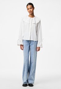 Weiße Bluse mit großem, gerüschtem Kragen, Bindeband am Hals, langen Ärmeln mit gerafften Manschetten; kombiniert mit hellblauen, weit geschnittenen Jeans.