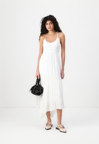White halter maxi dress with a textured bodice, floral lace accent, and tiered skirt. Paired with a black patterned handbag and shoes.