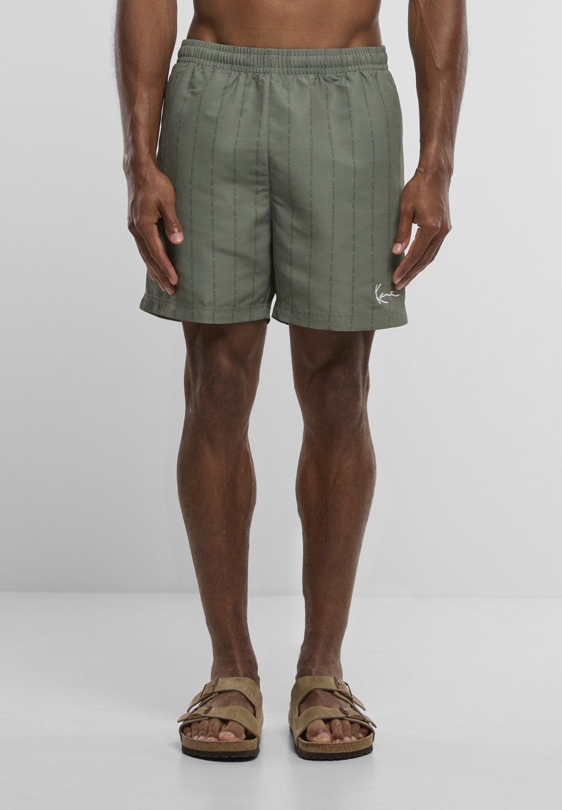 Modèle masculin portant un short de bain vert olive avec un motif de texte vertical subtil et des sandales à double bride couleur sable, debout sur un fond uni.