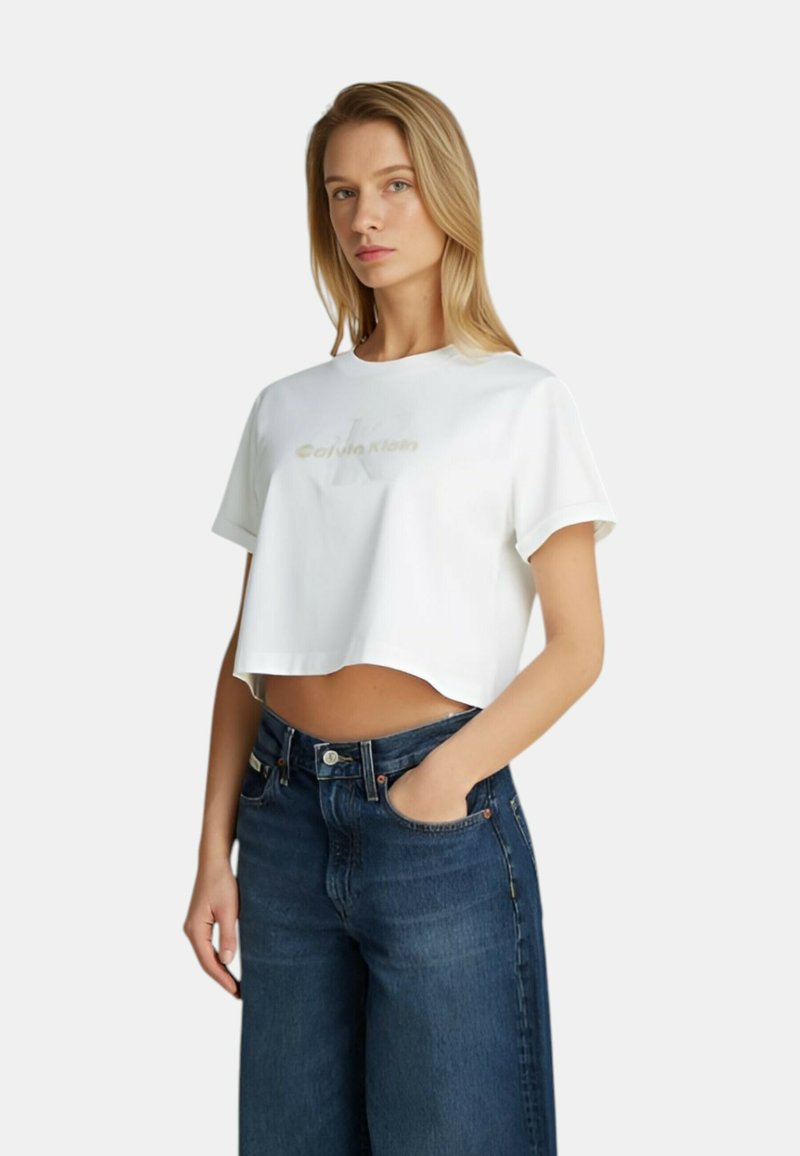 T-shirt bianca cropped in cotone con scollo rotondo e maniche corte, caratterizzata da un sottile logo beige di Calvin Klein sul davanti.