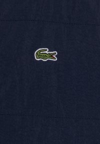 Un tissu bleu marine avec une finition texturée présente un petit logo de crocodile vert brodé. Design simple avec une couture droite au-dessus du logo.