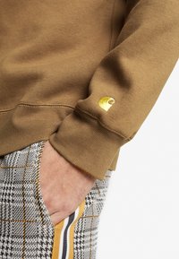 Brun sweatshirt med ribbade muddar och en guldfärgad logotyp. Matchad med mönstrade grå byxor med gula och svarta ränder längs sidorna.