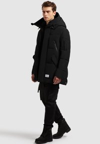 Juoda pūkinė striukė su aukštu apykakliu, šoniniais kišenėmis su užtrauktukais ir reguliuojama gaubtuvu. Derinama su juodomis kargo kelnėmis ir atitinkančiais batais.