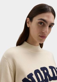 Suéter de lana crema con cuello alto y letras azules texturizadas que dicen "NORD". Puños y dobladillo acanalados, textura suave, diseño casual.