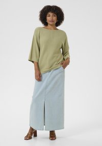 Hellgrüner Strickpullover mit weitem Ausschnitt, kombiniert mit einem hellblauen Jeansrock mit Schlitz vorne, ergänzt durch braune Riemchensandalen mit Absatz.
