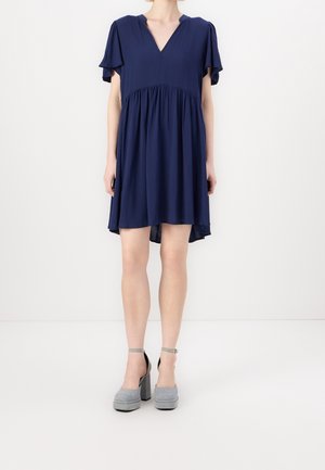 Vrouw draagt een losse marineblauwe korte jurk met wapperende mouwen en grijze platform-hakken, staand op een witte vloer.