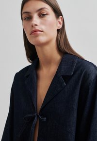 Jachetă din denim închis, cu guler, închidere cu cravată și mâneci largi. Prezintă o textură netedă și o nuanță de albastru închis.