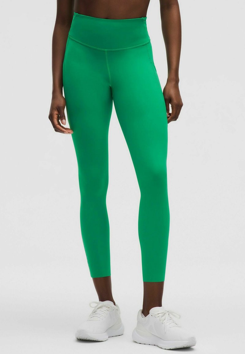 Zöld, magas derékú leggings, sima, rugalmas anyagból készült, varrás nélküli derékrésszel és testhezálló kialakítással. Fehér tornacipővel viselve.