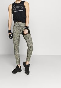 Svart ärmlös topp med vitt logo, mönstrade gröna leggings med svarta accenter, och svarta träningesskor, håller hantlar.