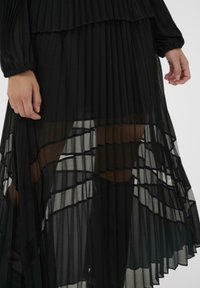 Čierna plisovaná sukňa s priehľadnými vrstvami, rôznou priehľadnosťou a elastickými manžetami. Detailná textúra s horizontálnymi a vertikálnymi líniami.