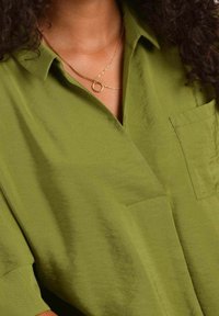 Green collared blouse with a textured finish, featuring a chest pocket and short sleeves, worn with a layered gold necklace.