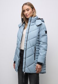 Vrouw draagt een lichtblauwe doorgestikte winterjas met capuchon, staat met haar handen langs haar zij tegen een effen witte achtergrond.